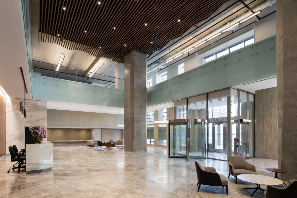 EQ Bank in Toronto, Canada atrium featuring a custom LINE:Form Wave Baffle ceiling feature in extruded metal that flows overhead. The ceiling’s undulating form brings a dynamic sense of movement and architectural expression to the open space. Finished in a warm wood veneer, the baffles offer a material counterpoint to the exposed cement walls, granite flooring, and structural pillars, softening the palette while maintaining a refined, modern aesthetic.